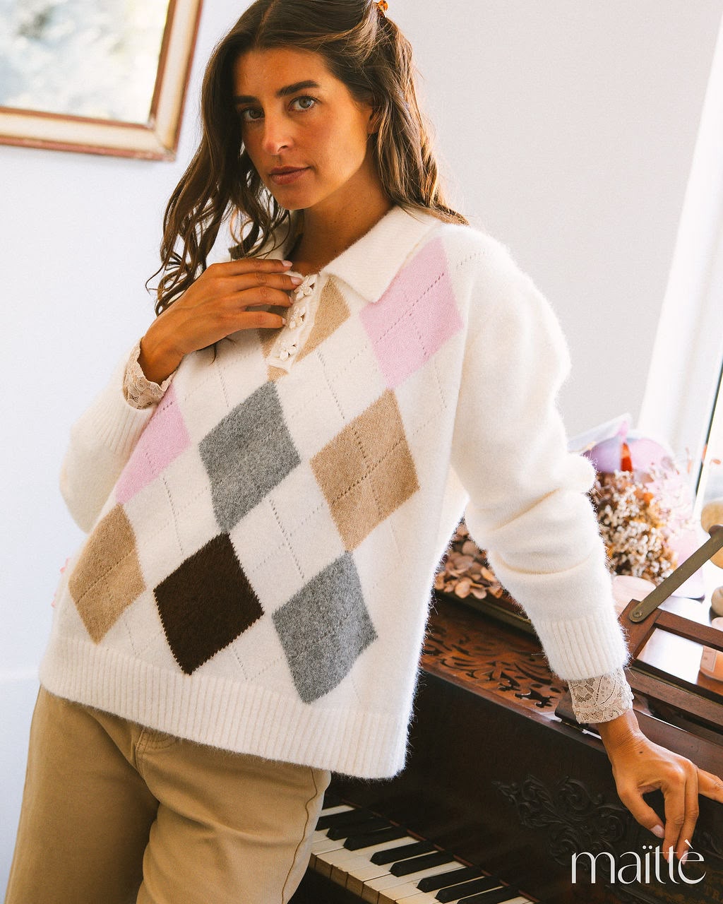 CONJUNTO CIANA Mulher com camisola de malha branca com padrão argyle colorido encostada a um piano de madeira escura