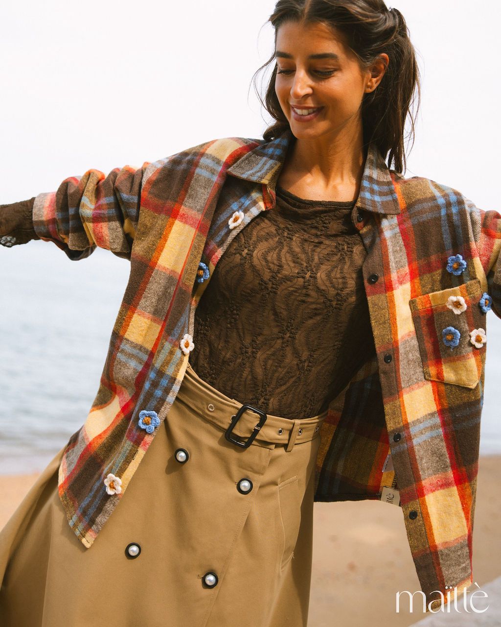 CONJUNTO CASTELINA Mulher com casaco xadrez colorido, blusa rendada castanha e saia bege na praia