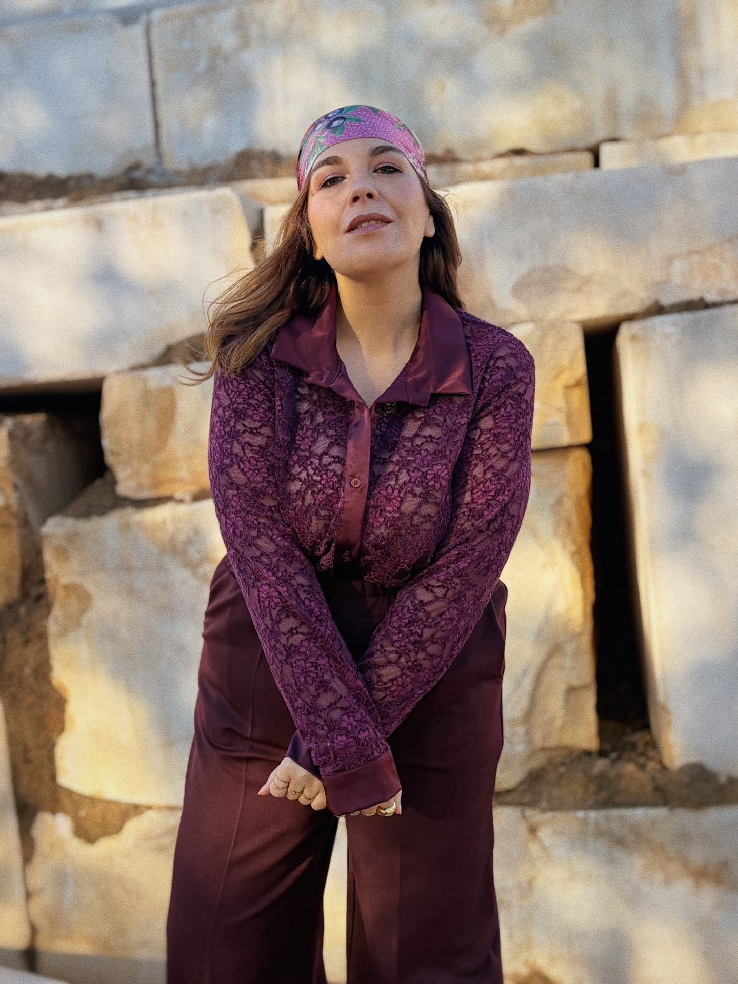 CAMISA ANDREIA Mulher com lenço rosa, blusa de renda roxa e calças roxas em fundo de pedras