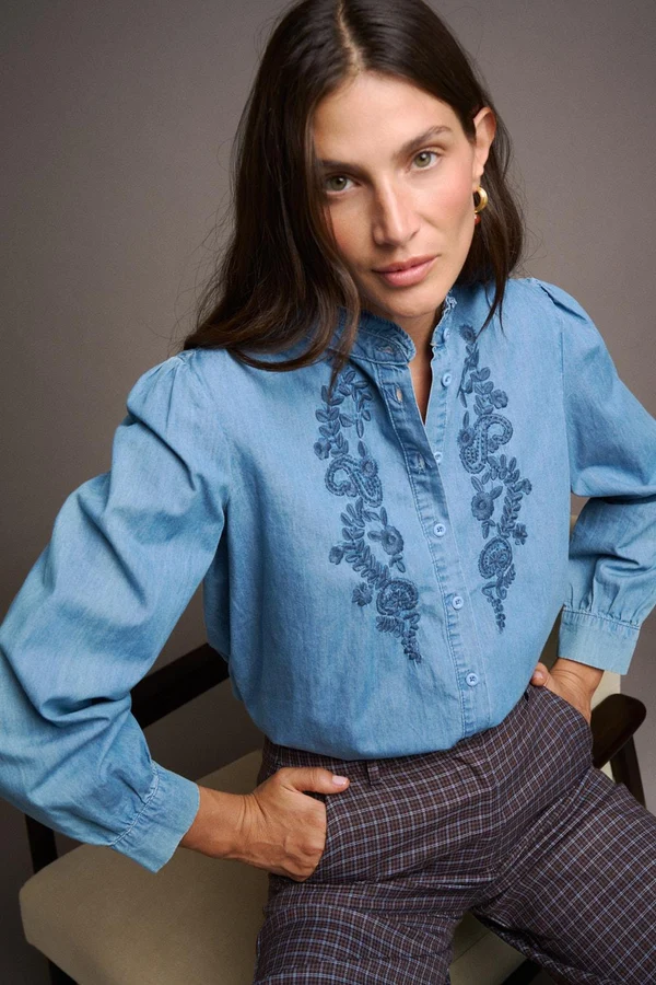 BLUSA MIRANDA Blusa azul clara com bordado floral e calças xadrez castanhas usadas por mulher sentada