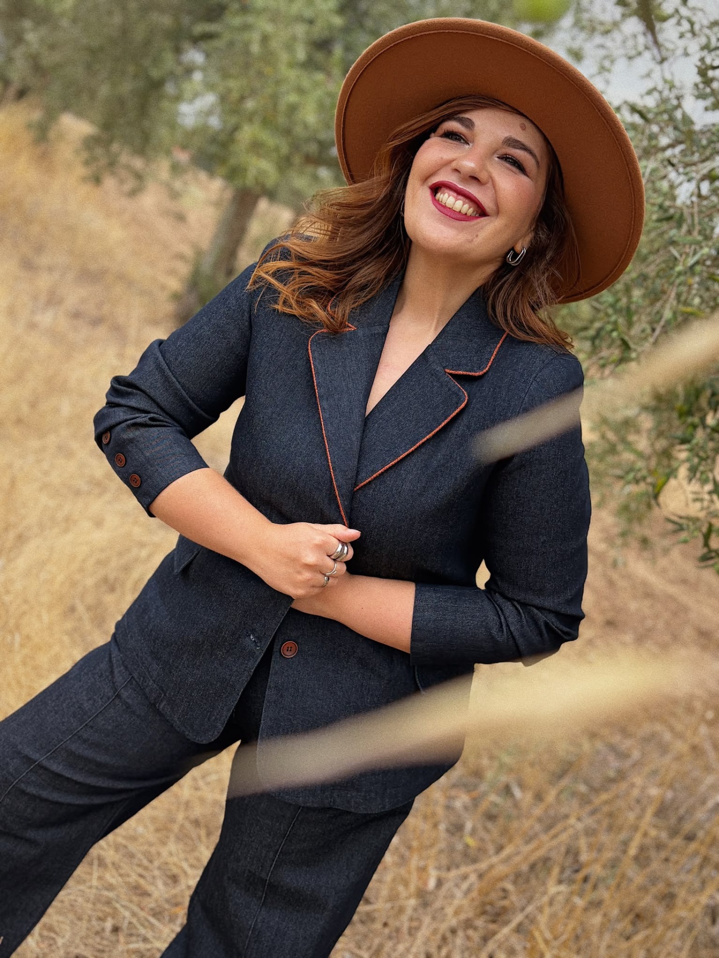 BLAZER AMÉLIA Mulher sorridente com blazer cinza escuro com costura laranja, calças cinza escuro e chapéu castanho em ambiente natural