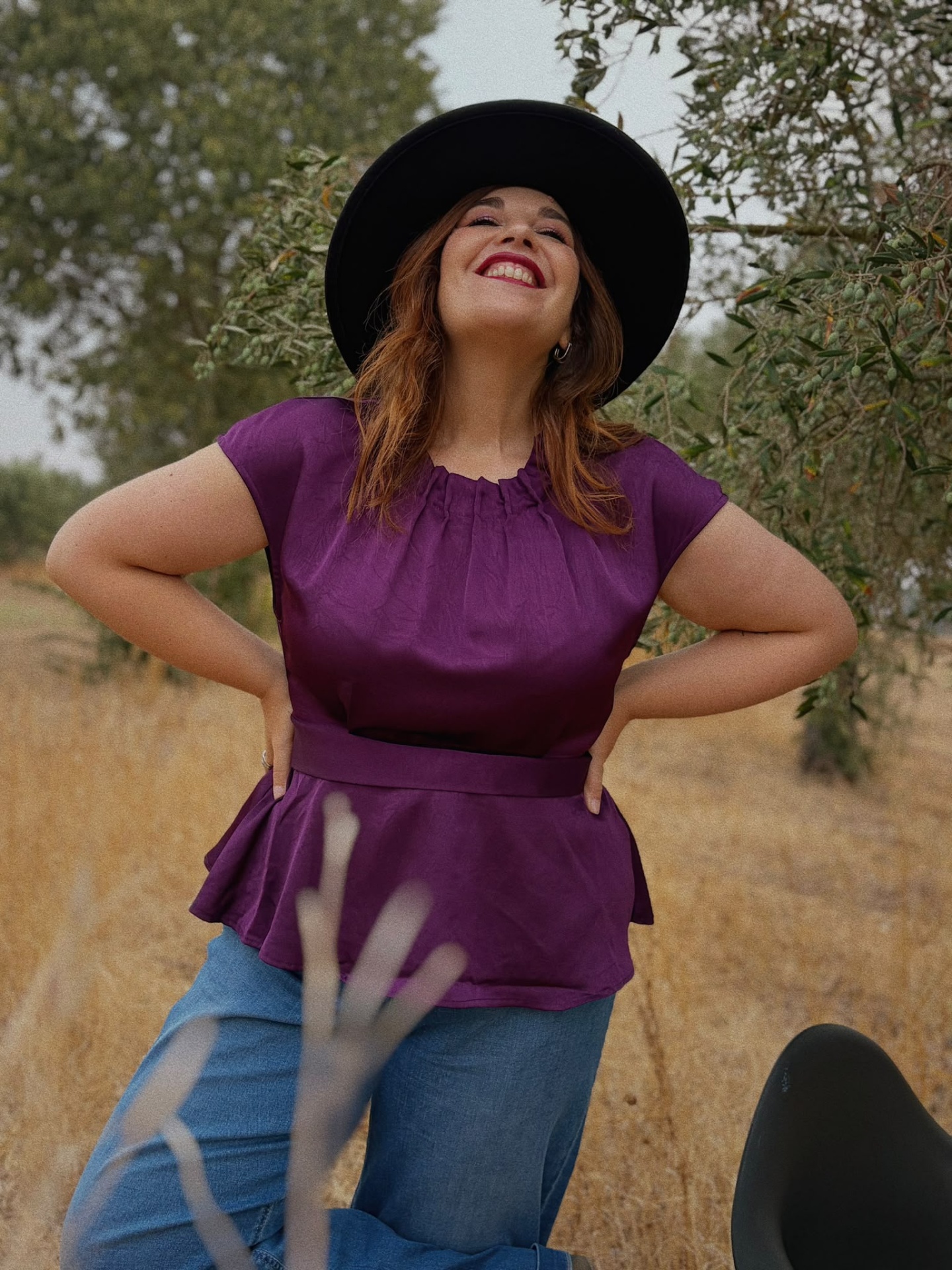 TOP AMABÉLIA BERINGELA Mulher sorridente com blusa roxa e chapéu preto ao ar livre