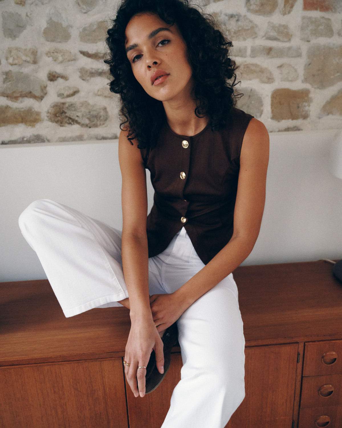 CALÇAS RAVEN BRANCO Mulher com top castanho e calças brancas sentada numa cómoda de madeira com parede de pedra ao fundo