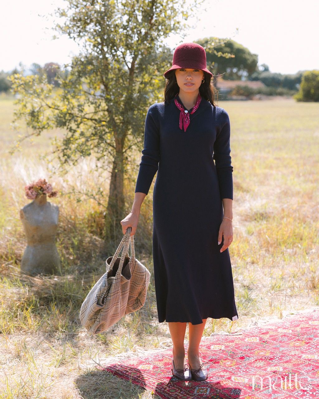 VESTIDO ARTEMISA Mulher com vestido azul escuro, cachecol vermelho, chapéu vinho e bolsa xadrez em campo com tapete vermelho.