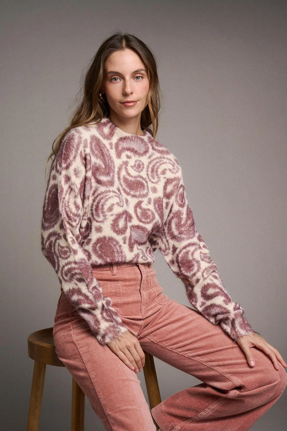 CAMISOLA MERCEDES Mulher com camisola de padrão paisley creme e rosa e calças cor de rosa sentada num banco de madeira.