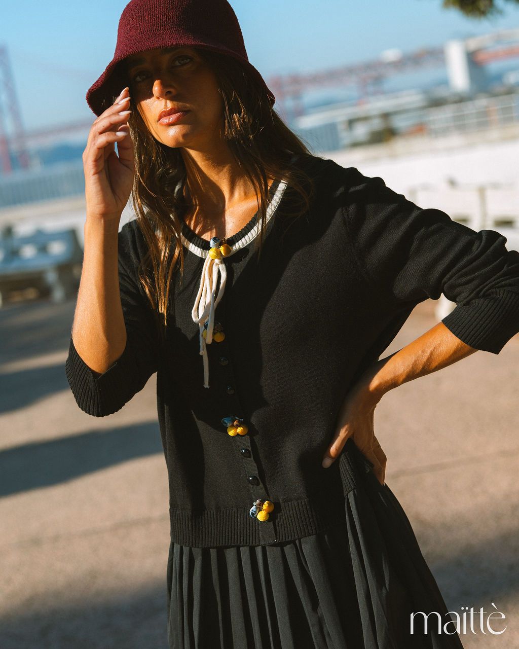 VESTIDO BASÍLIA Mulher com cardigan preto com botões decorativos, vestido preto plissado e chapéu vinho ao ar livre