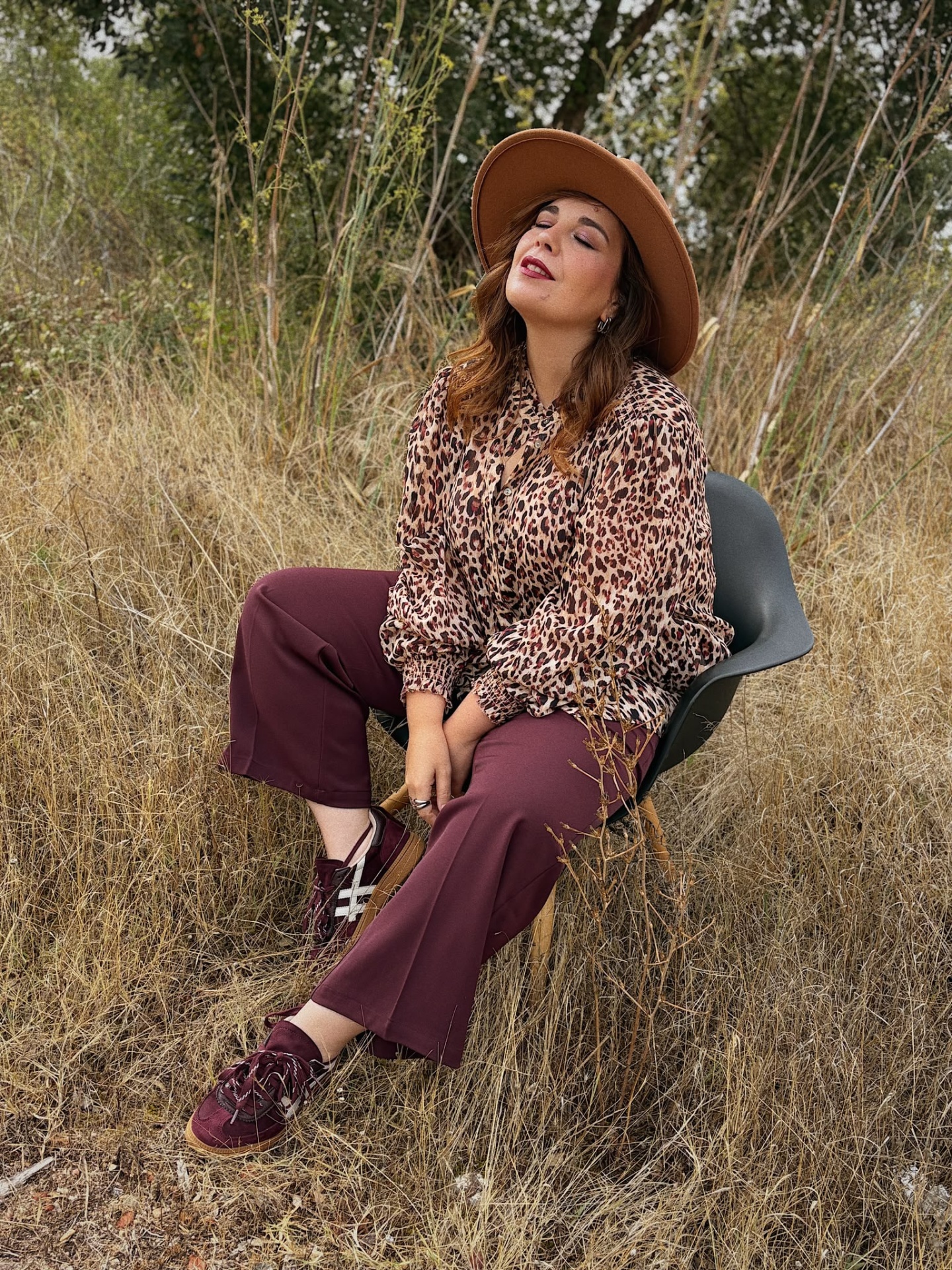 CALÇAS NOBLESSE VINHO Pessoa sentada em cadeira ao ar livre com blusa padrão oncinha, calças bordô, boné castanho e sapatilhas bordô
