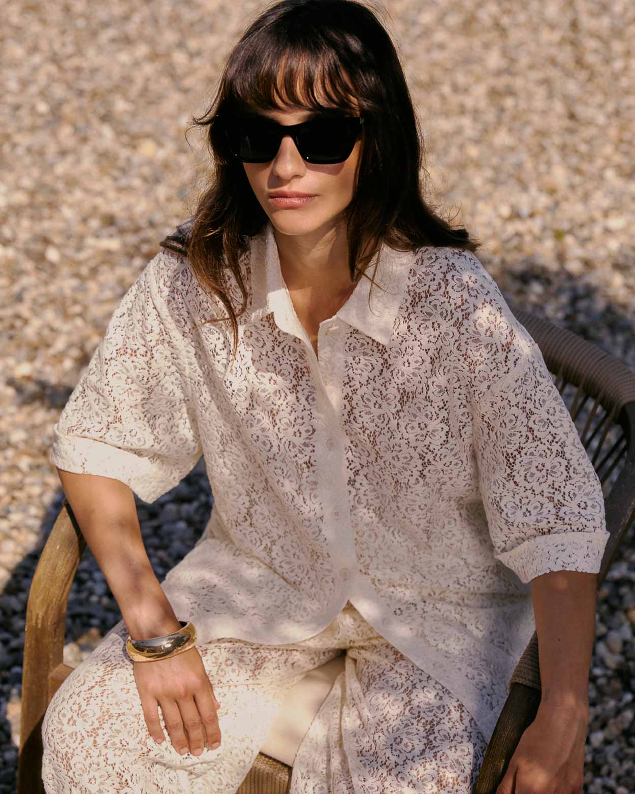 CAMISA VENDEL ECRU Mulher de blusa e calças em renda bege com padrão floral, óculos escuros pretos, sentada em cadeira de madeira sobre chão de pedras