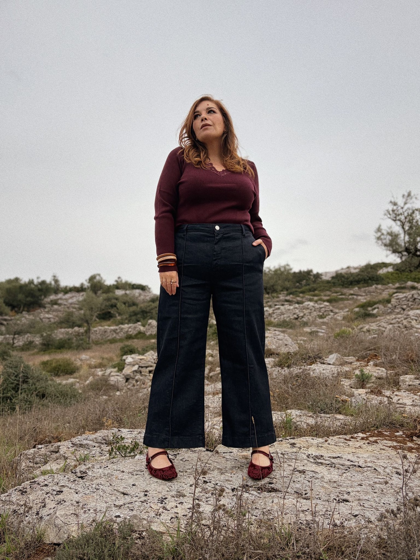 CALÇAS AMINATA Mulher com camisola bordeaux, calças pretas largas e sapatilhas bordô numa paisagem rural rochosa.