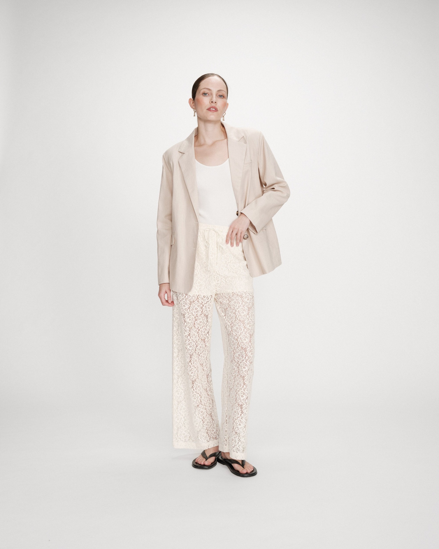 CALÇAS VICENT ECRU Mulher com blazer bege, top branco, calças rendadas brancas e sandálias pretas