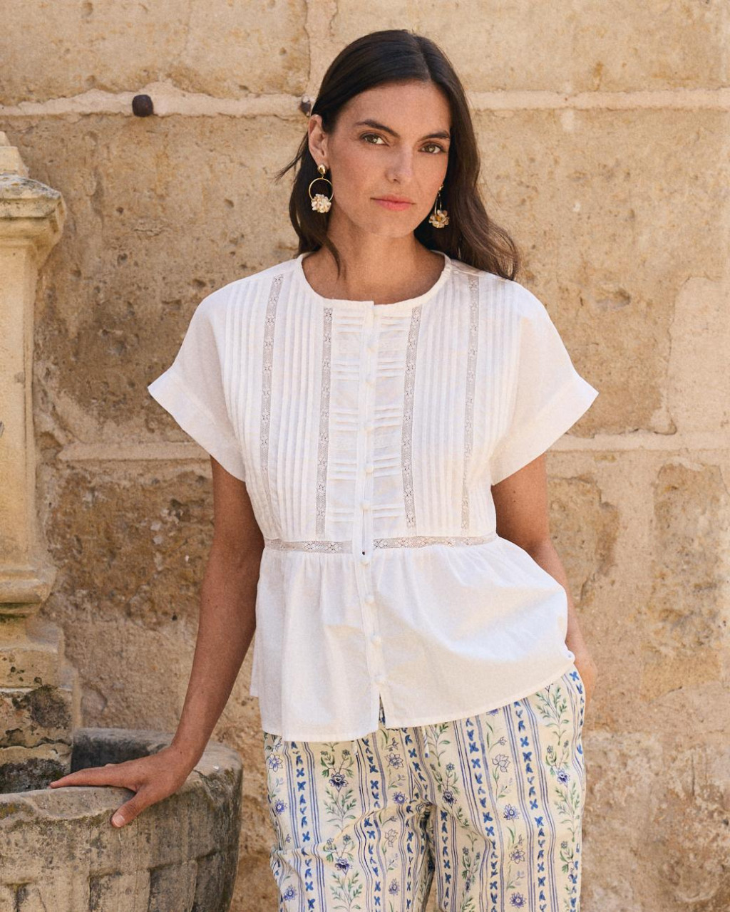 BLUSA BLANCA MAITE Mulher com blusa branca de manga curta e calças florais perto de parede de pedra