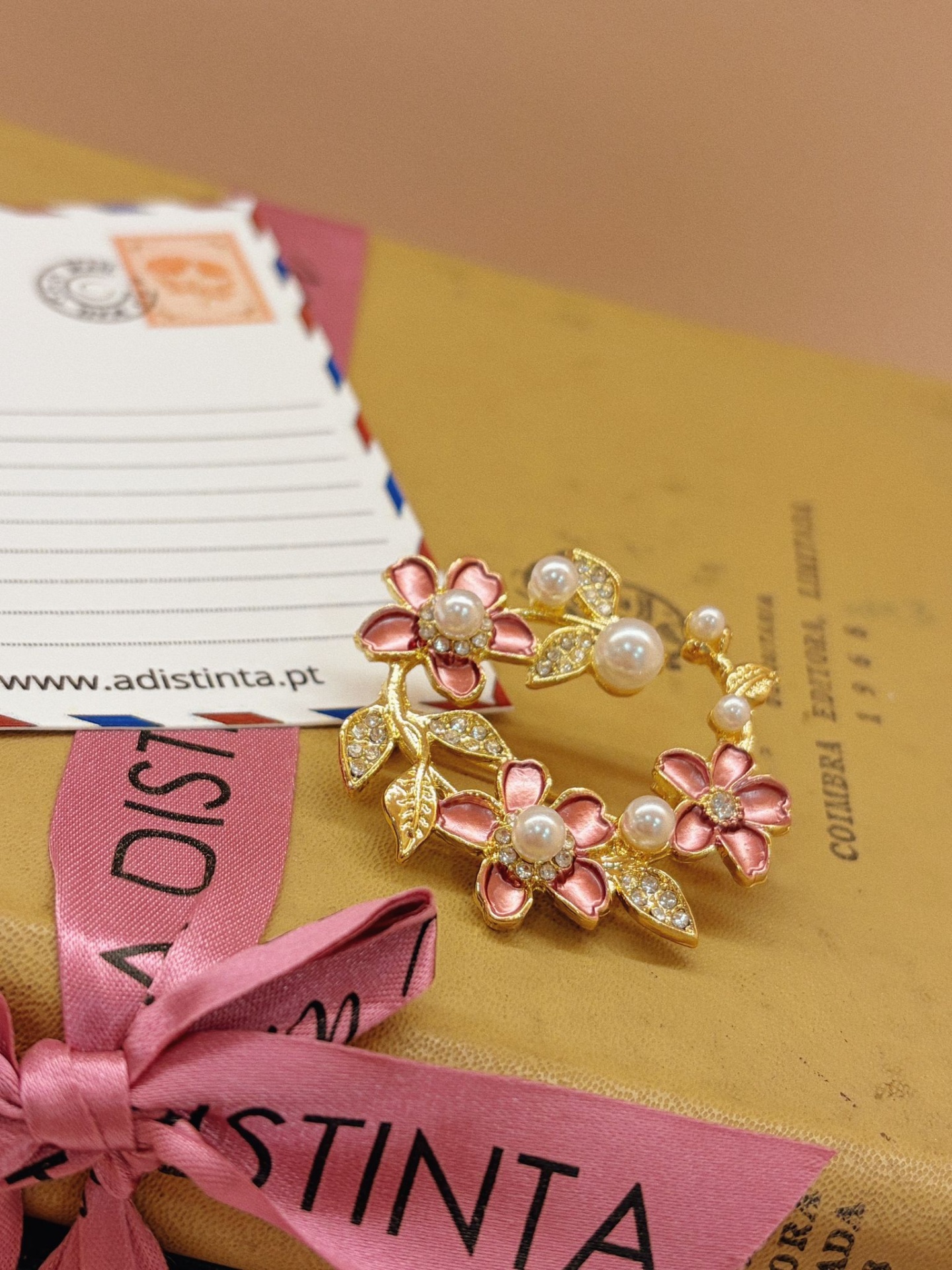 PREGADEIRA QUAR Anel dourado com flores cor de rosa, pérolas e folhas com cristais sobre caixa amarela e laço rosa