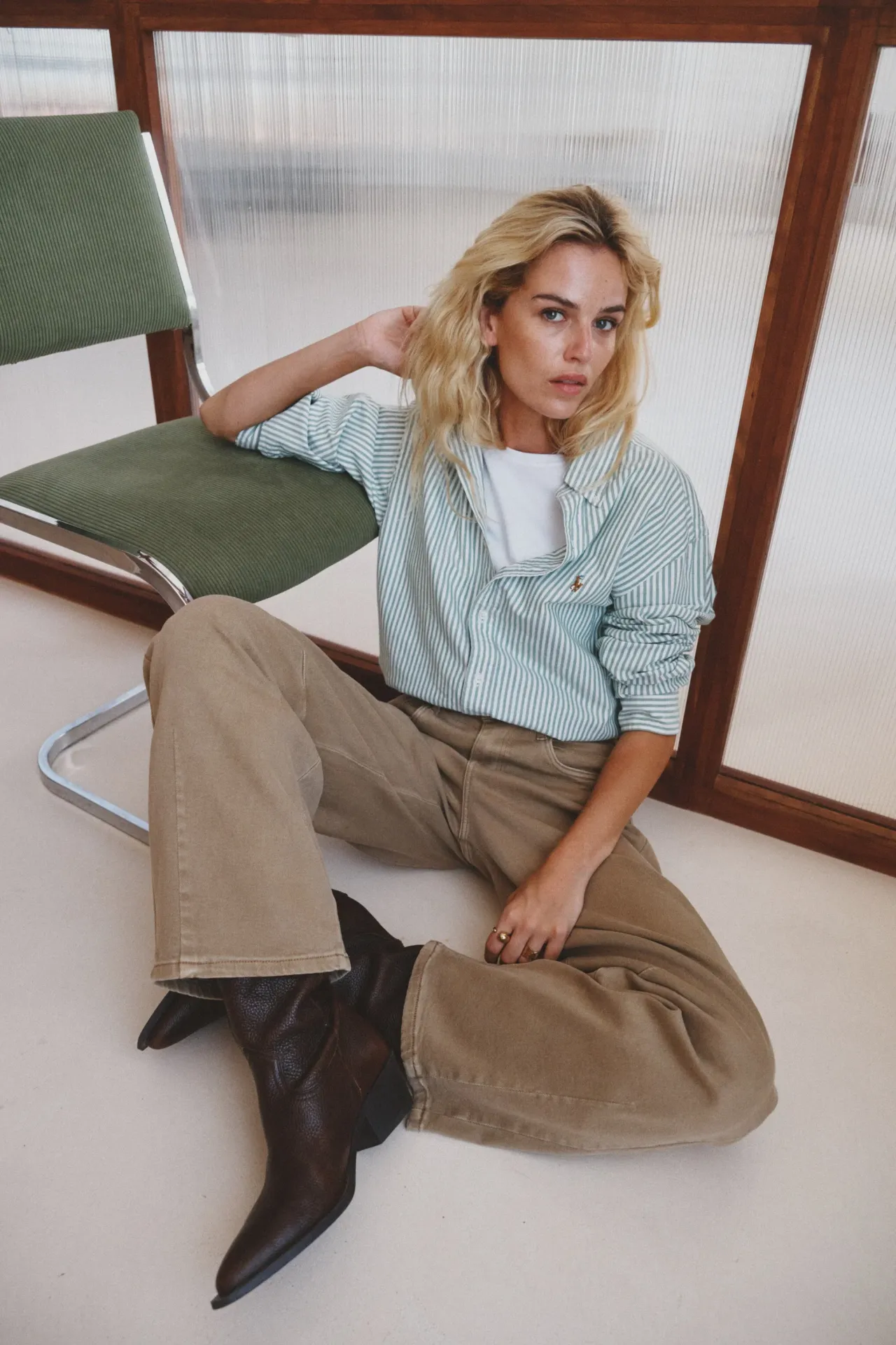 CALÇAS BARREL CREME Mulher sentada no chão com camisa às riscas verde claro e branco, calças bege largas e botas castanhas, ao lado de cadeira verde com estrutura metálica.
