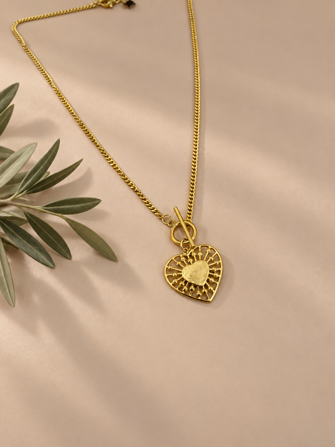 FIO BERENGÁRIA Colar dourado com pendente em forma de coração e corrente sobre fundo bege com folhas verdes ao lado