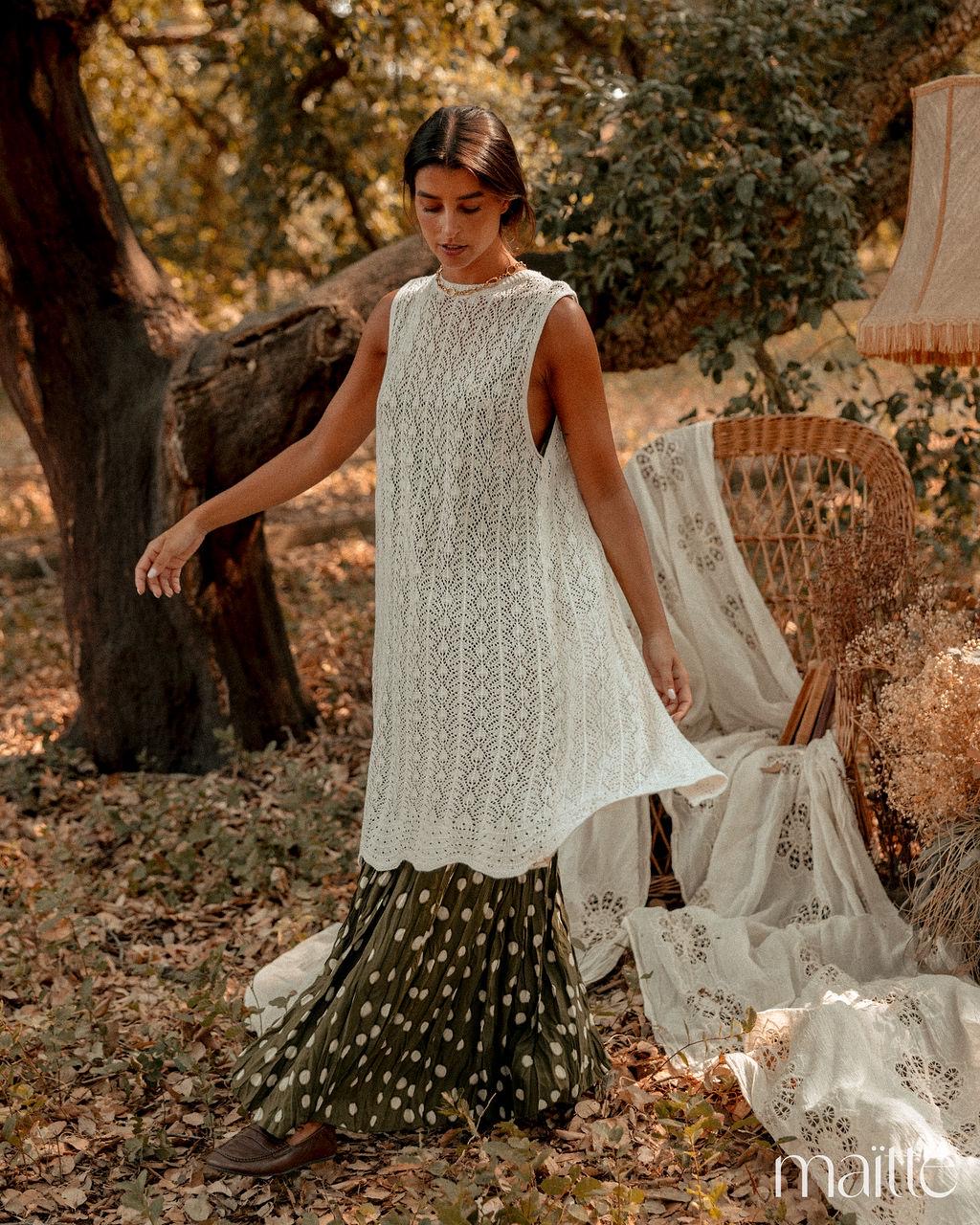 CONJUNTO ANGÉLICA Mulher com vestido branco com padrão de renda e saia verde com bolinhas brancas em ambiente exterior com folhas secas e cadeira de vime