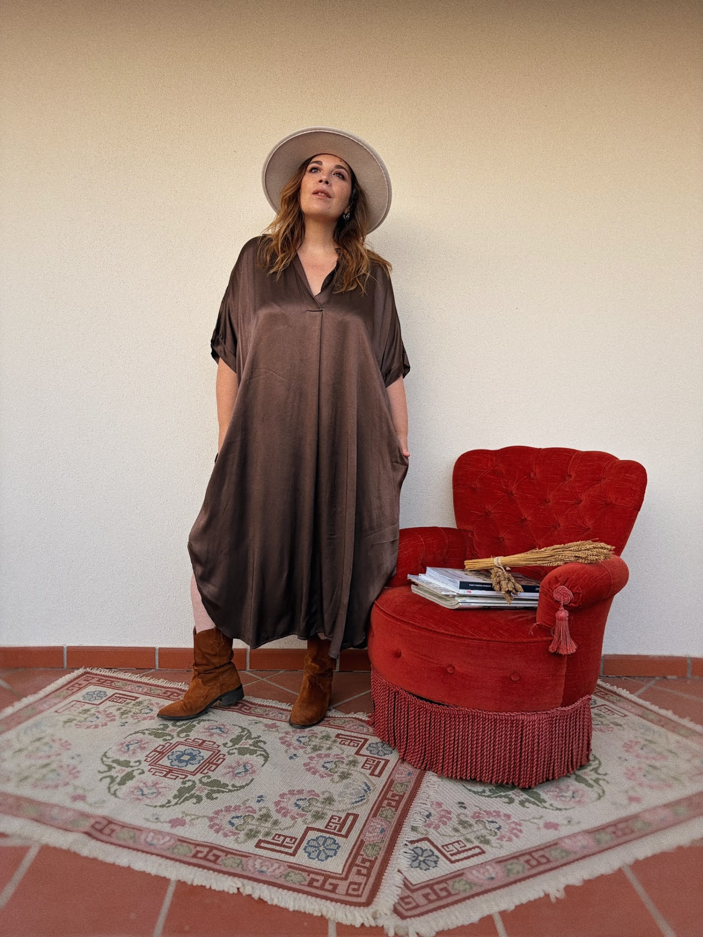 VESTIDO ALMA Mulher de vestido castanho fluído, chapéu branco, botas castanhas, ao lado de poltrona vermelha e livros.