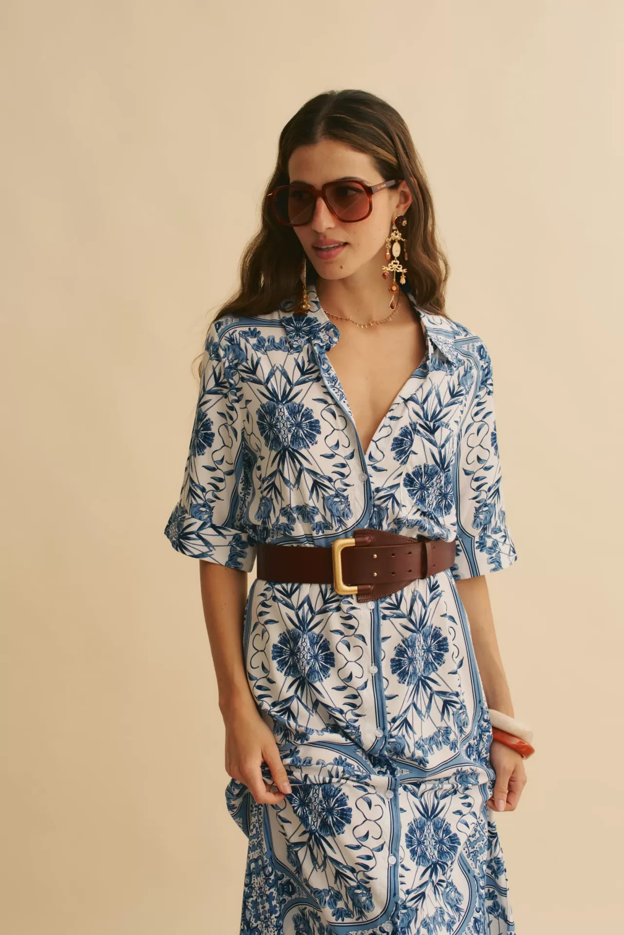 VESTIDO DENIM DREAMS CAMISEIRO Mulher com vestido branco e azul, cinto castanho, óculos de sol e brincos grandes
