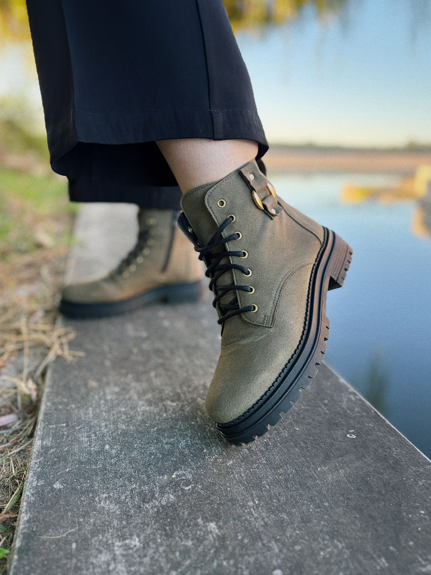 BOTAS CAROL KHAKI Bota verde-oliva de camurça com sola grossa e fivela dourada, usada com calças pretas perto de água.