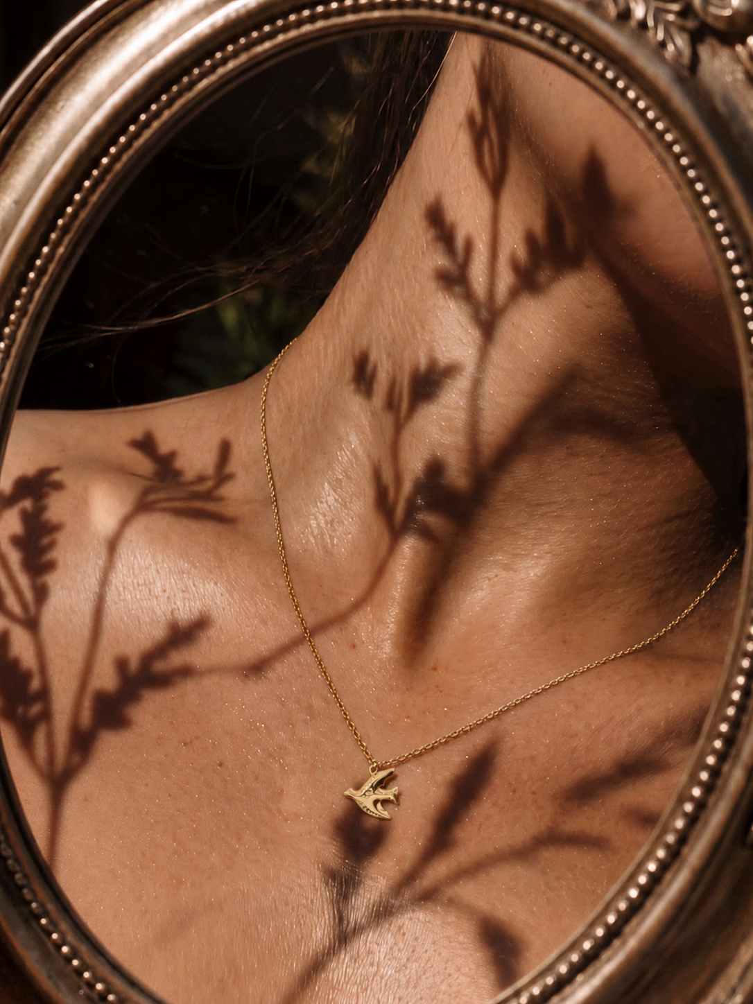 FIO ANAÍS Colar dourado com pendente de pássaro e sombras de ramos no pescoço refletido em espelho antigo.