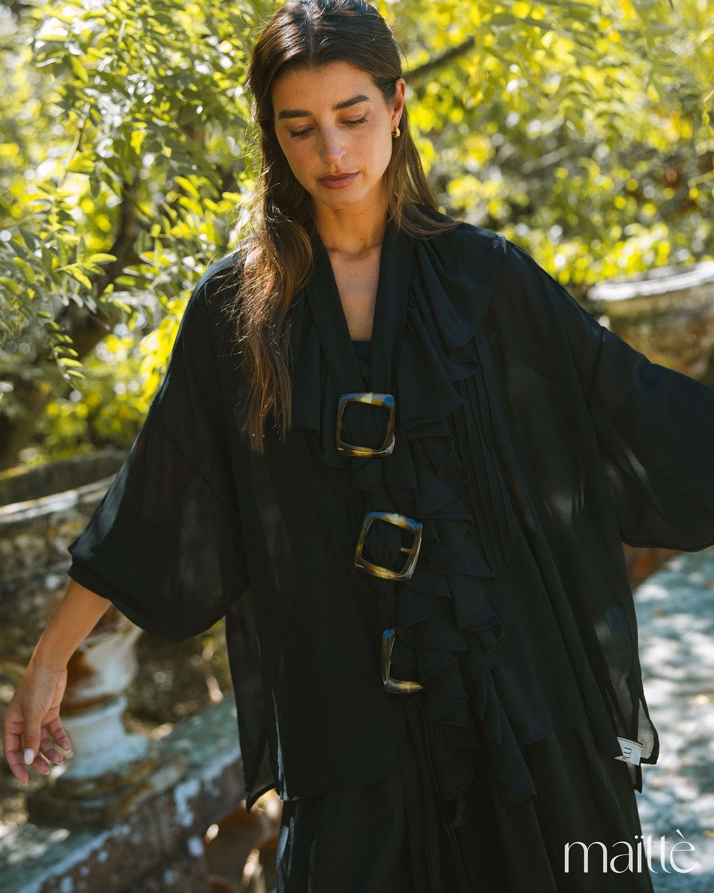 TÚNICA CANDICE Mulher a usar top preto fluído com folhos e fivelas grandes em ambiente exterior verde