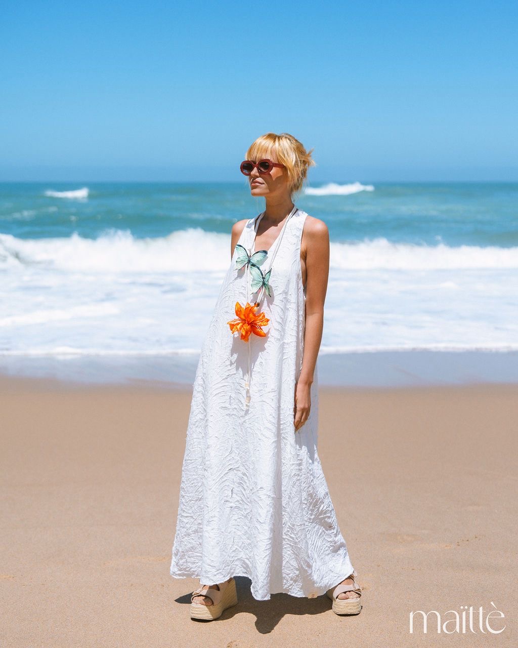 VESTIDO ZENAIDA mulher com vestido longo branco e colar de flores na praia