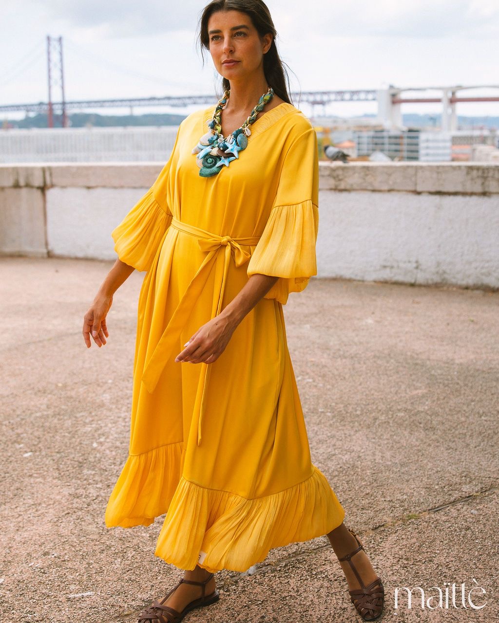VESTIDO VLADA Mulher com vestido amarelo comprido e colar grande colorido em ambiente exterior com uma ponte ao fundo.