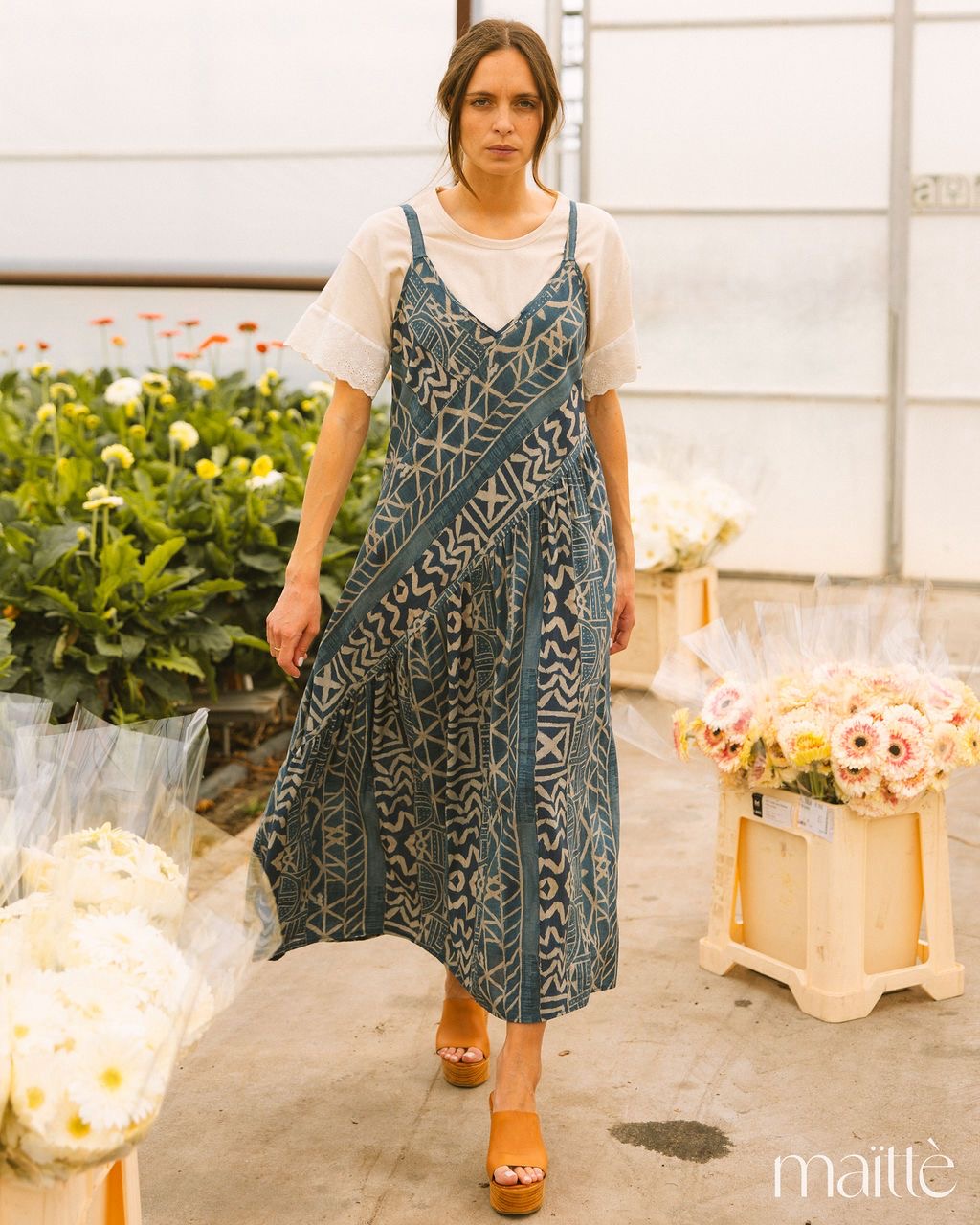 VESTIDO ZARINA Mulher com vestido comprido azul com padrões geométricos a usar t-shirt branca e sandálias castanhas, num local com flores e caixas de plástico.