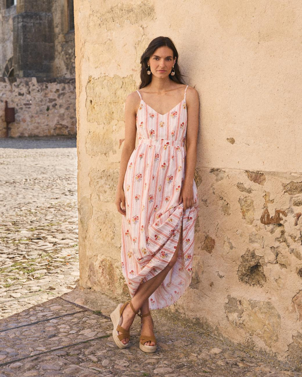 VESTIDO NATALIA Mulher com vestido comprido listrado rosa com flores encostada numa parede de pedra
