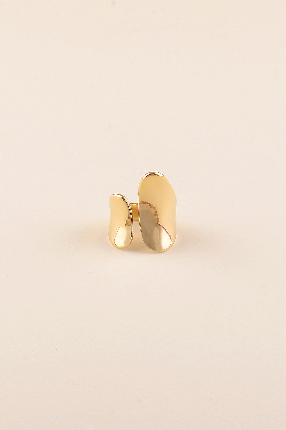 ANEL SHAPES Anel dourado com duas formas orgânicas lisas e polidas, sobre fundo bege claro.