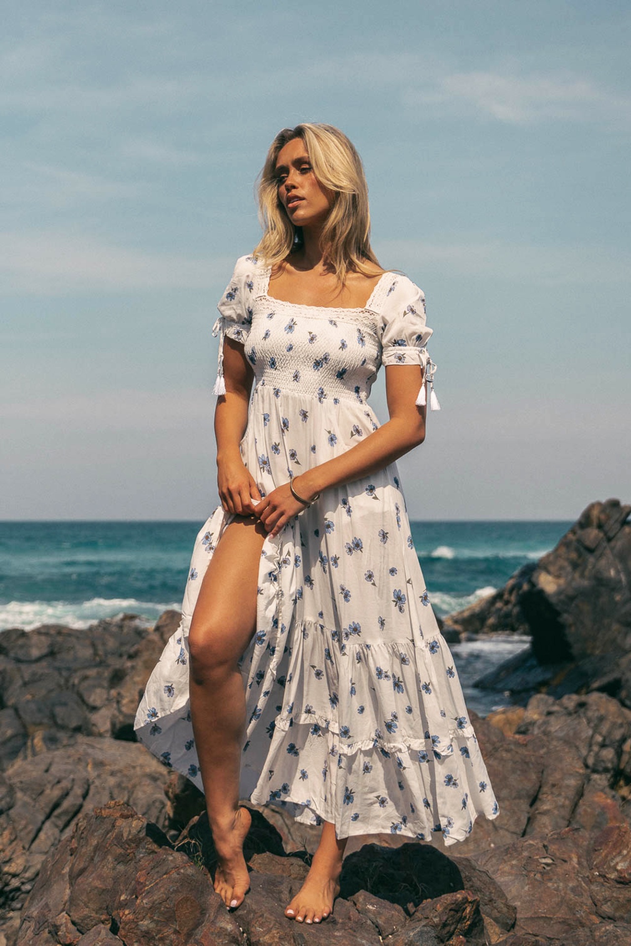 VESTIDO JUNEBERRY Mulher com vestido longo branco floral em praia rochosa.