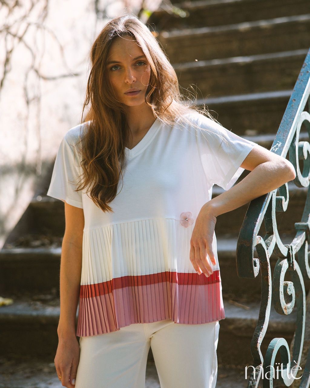 T-SHIRT SUATI Camisola branca com detalhe plissado e flor rosa, usada por mulher, em escadas exteriores com grade azul