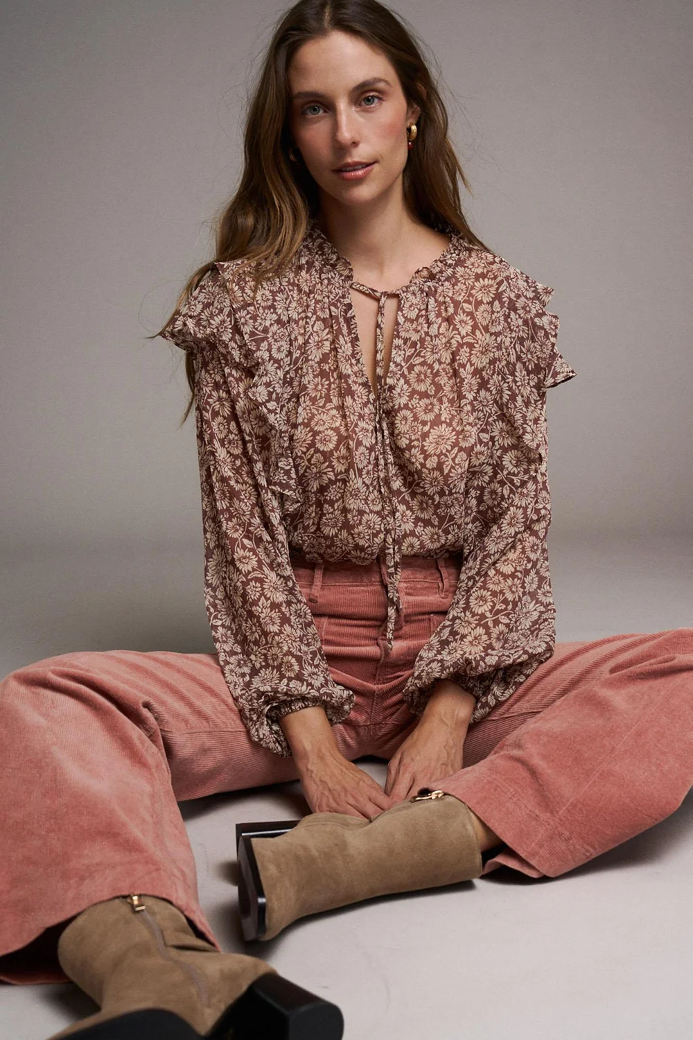 BLUSA DÁCIL Mulher sentada a usar blusa floral com folhos, calças rosa de veludo cotelê e botas castanhas.