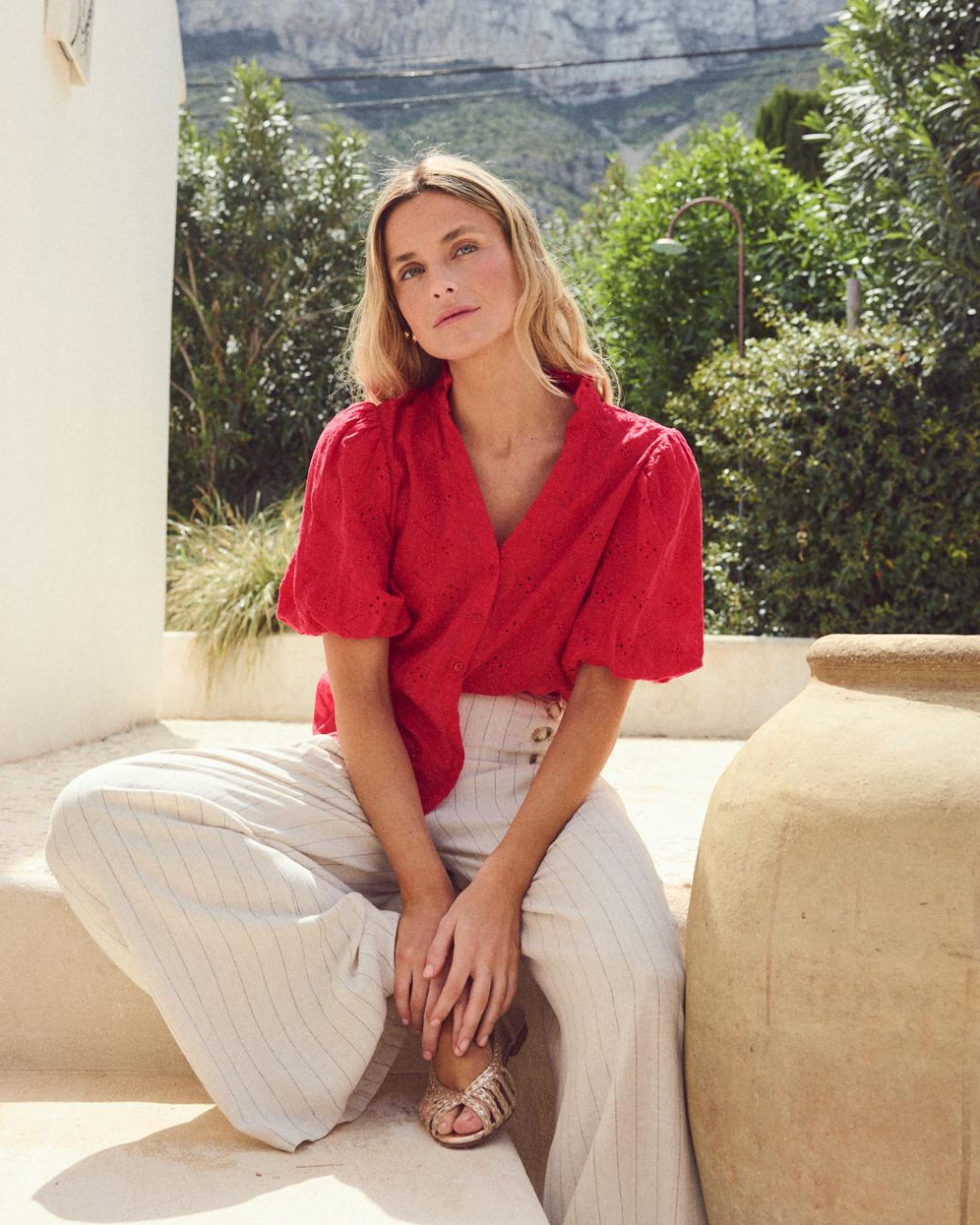 BLUSA GEMA Mulher com blusa vermelha e calças largas bege num cenário exterior com plantas e vaso cerâmico.