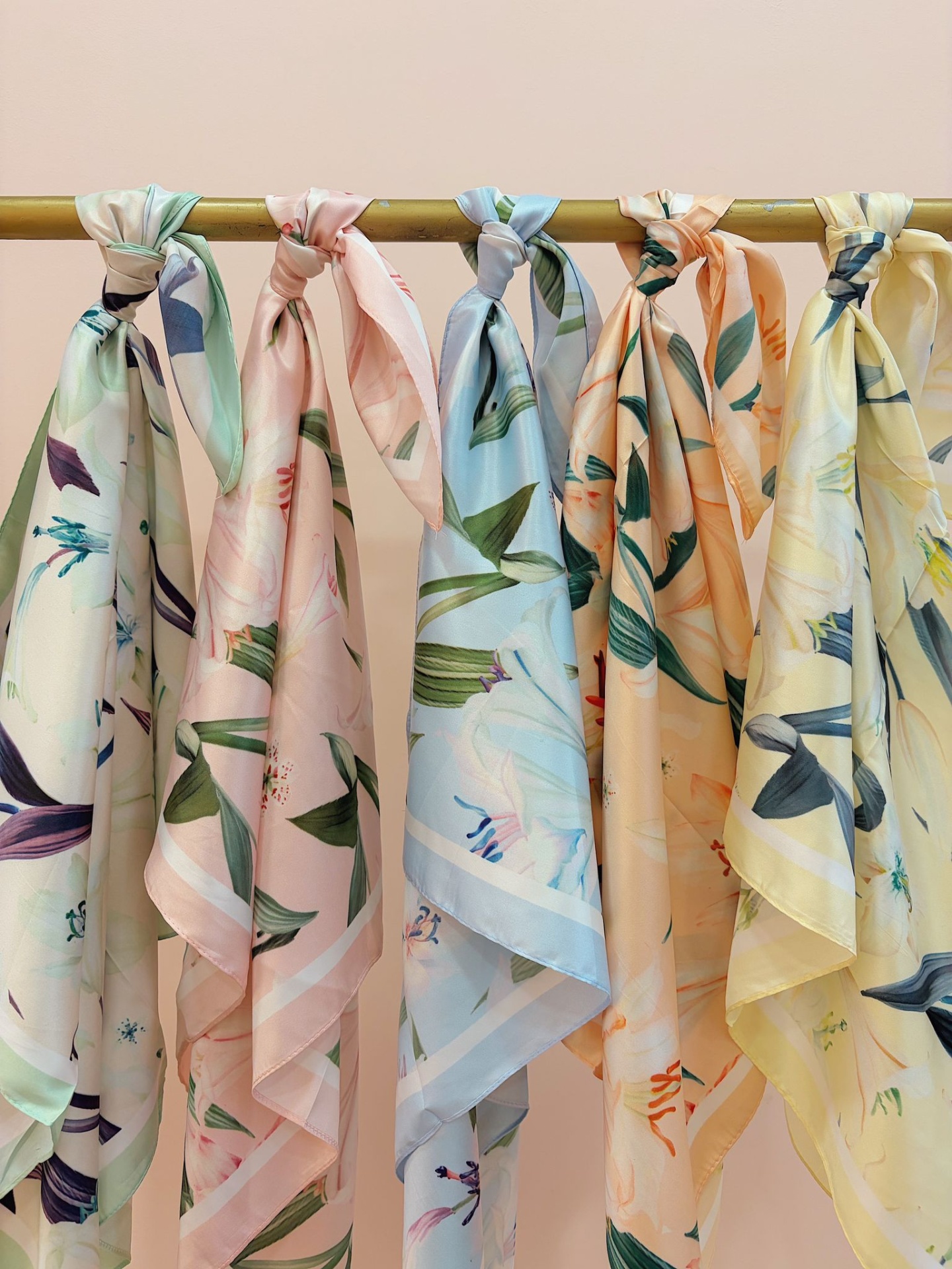LENÇO STANISLAVA Seis lenços de padrão floral em cores pastel pendurados numa barra metálica dourada.