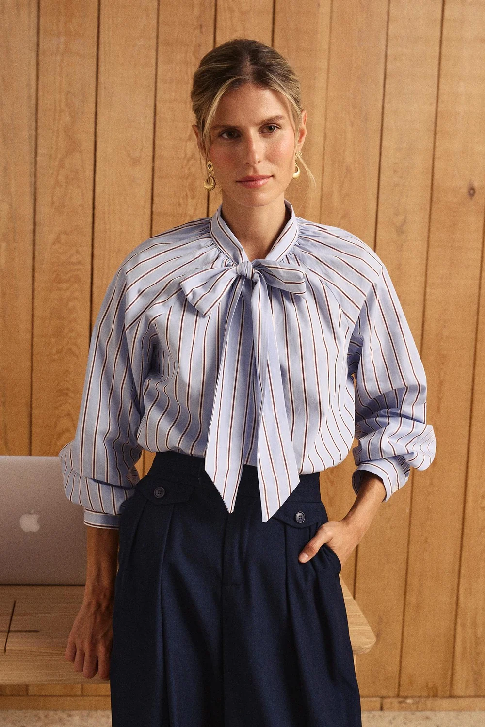 BLUSA GIOMAR Mulher com camisa de riscas azul clara com laço e calças azul-escuras em ambiente de madeira
