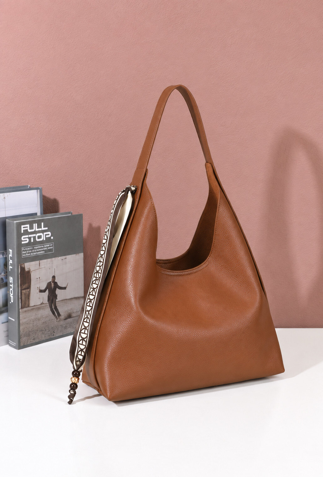 MALA BRÍGIDA Mala castanha de mão com fita decorativa pendurada e livros ao fundo sobre mesa branca