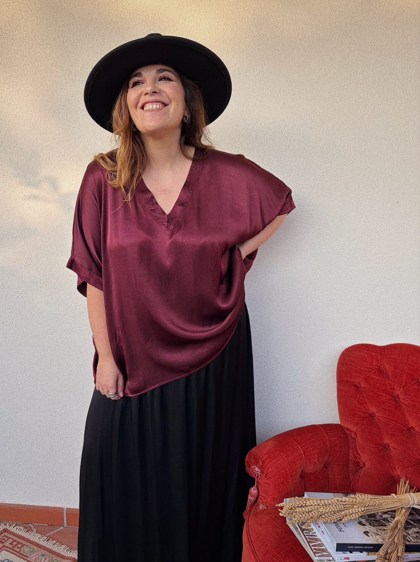 BLUSA ALMARA Mulher com blusa vinho acetinada, saia preta e chapéu preto em ambiente interior junto a cadeira vermelha