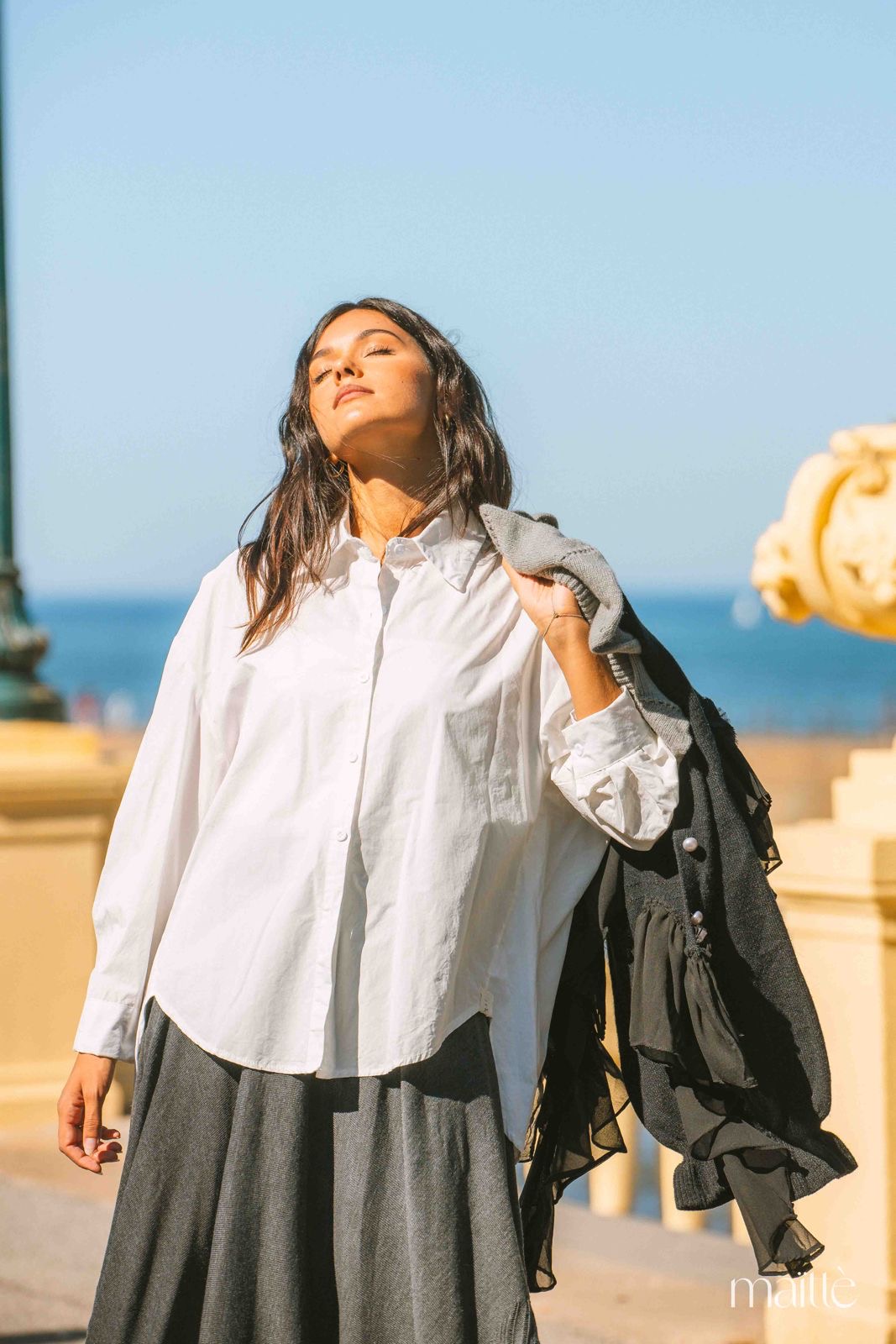 CONJUNTO CARMELINDA Mulher com camisa branca, saia cinza e casaco preto ao ar livre com céu e mar ao fundo.