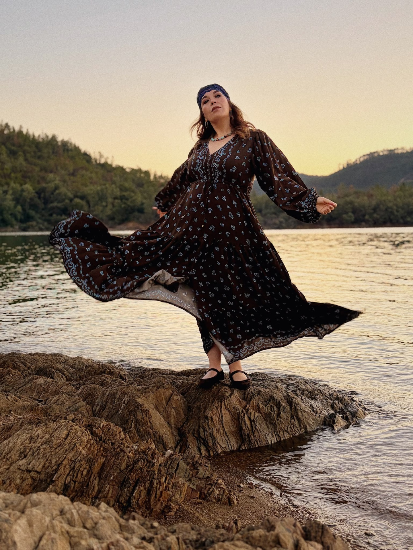 VESTIDO JUNIPER Mulher com vestido comprido castanho de flores azuis junto a lago e colinas ao pôr do sol