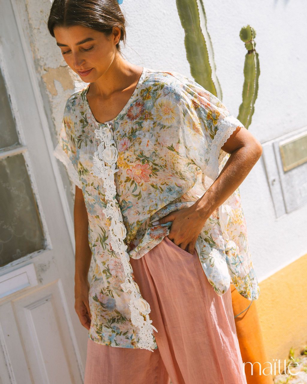 CAMISA ABNA Mulher a usar blusa floral com renda branca e calças largas cor de rosa num ambiente exterior urbano.