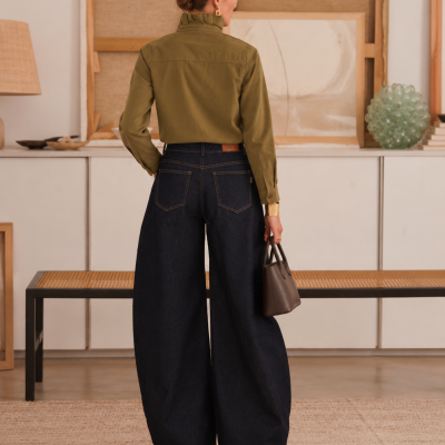 Mulher vista de costas com calças largas de ganga azul escura e camisa verde, segurando mala castanha em ambiente interior