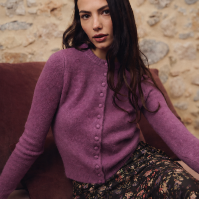 Mulher com casaco roxo e saia floral sentada num sofá com almofadas castanhas.