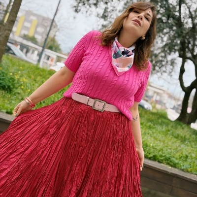 Mulher com camisola rosa, saia vermelha plissada, cinto rosa, lenço colorido e pulseiras, em parque exterior.