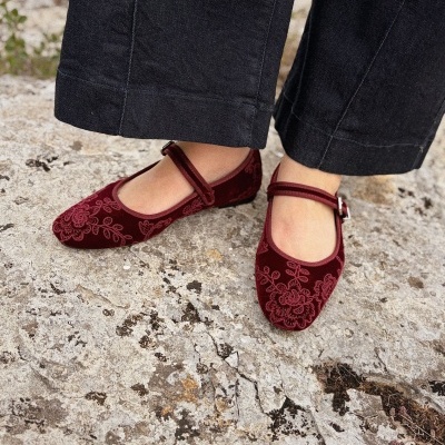 Sapatos Mary Jane vermelhos em veludo bordado usados com calças pretas sobre chão de pedra.