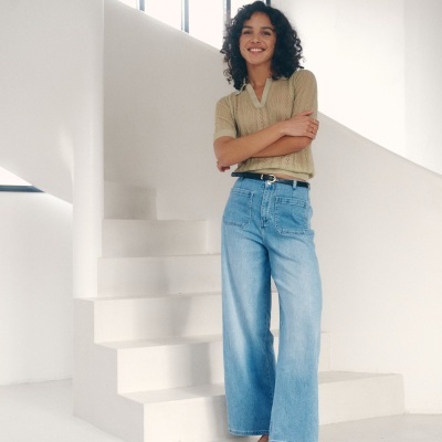 Mulher com camisola bege e jeans de ganga azul claro largas, descalça, em interior branco moderno com escada