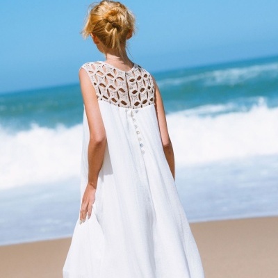 Vestido branco comprido com costas em renda branca e botões decorativos, vista de costas, praia ao fundo.