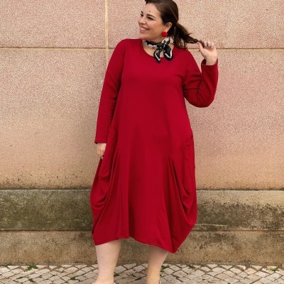 Mulher de vestido vermelho com bolsos grandes, lenço no pescoço e sapatilhas bege frente a parede rosa claro