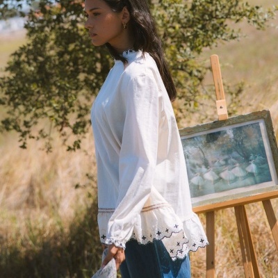 Blusa branca com renda e bordados, usada por mulher ao ar livre perto de pintura em cavalete