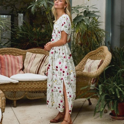 Mulher com vestido comprido branco estampado com flores e sandálias castanhas, em ambiente interior com móveis de vime e plantas