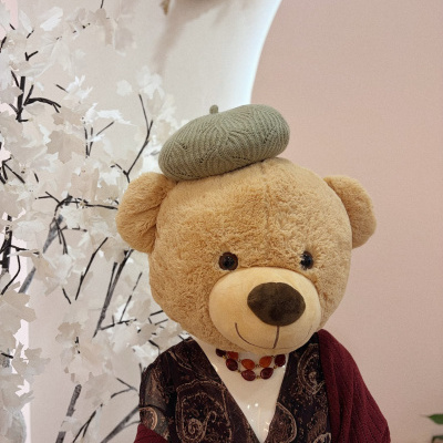 Boneco de peluche urso com barretina verde e roupa padrão castanho e vermelho num cenário interior com parede rosa e planta branca